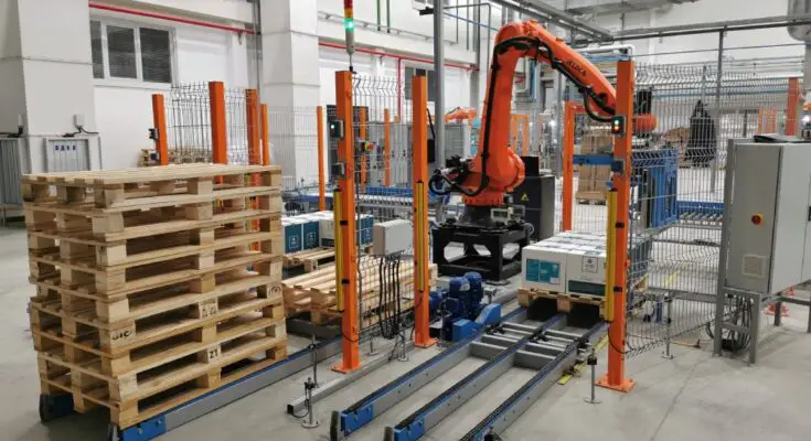 An orange palletizer machine stacks white cardboard boxes on a wooden pallet. A pile of empty pallets is next to the machine.