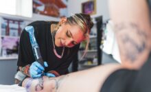 A female tattoo artist in blue gloves using a large needle covered in plastic to draw a tattoo on an individual's leg.