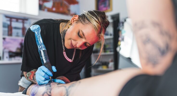A female tattoo artist in blue gloves using a large needle covered in plastic to draw a tattoo on an individual's leg.