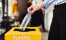 A woman in professional clothing discarding a plastic bottle into a yellow bin with "Plastic" written on the lid.