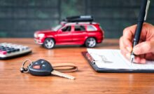 A person uses a pen to write on a piece of paper clipped to a clipboard. A toy car and set of keys sit on the table.