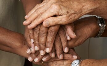 A close-up view shows three pairs of hands stacked on top of each other as a sign of support. Two wrists have watches.