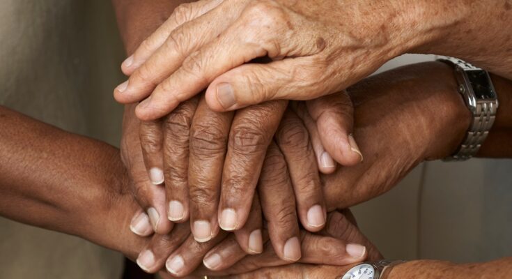 A close-up view shows three pairs of hands stacked on top of each other as a sign of support. Two wrists have watches.