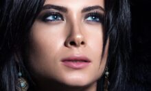 A close-up view shows a woman with dark hair and blue eyes. She's wearing brown feather earrings.