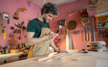 A man stands in his carpenter's workshop. He has pieces of scrap wood in front of him as he scrolls on his phone.