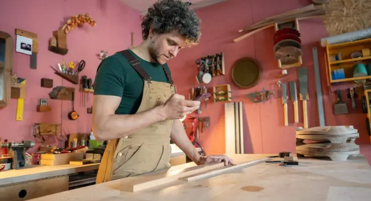 A man stands in his carpenter's workshop. He has pieces of scrap wood in front of him as he scrolls on his phone.