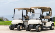 Two white golf carts with beige tops parked next to each other on a concrete road with trees in the distance.
