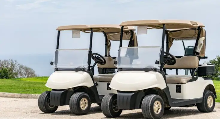 Two white golf carts with beige tops parked next to each other on a concrete road with trees in the distance.