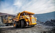A large yellow mining truck sitting in a mining quarry with hills and another piece of mining equipment behind it.