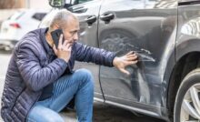 A man on his smartphone crouches down, placing his hand on the scratches and dents left on his gray sedan.