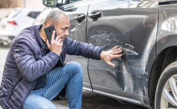 A man on his smartphone crouches down, placing his hand on the scratches and dents left on his gray sedan.