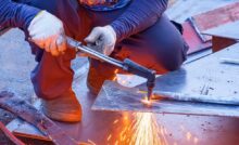 A person in long sleeves, pants, and gloves crouches while cutting an iron plate with an acetylene gas welding torch.