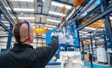 A worker points in the direction of an industrial crane as it moves a machine inside a large warehouse.