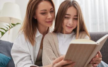 A close-up of a mom and teen daughter sitting together and reading a book on a couch. They're both smiling.