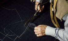 A close-up of a tailor in a striped shirt using a large pair of scissors to cut a pattern out of dark fabric.