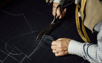 A close-up of a tailor in a striped shirt using a large pair of scissors to cut a pattern out of dark fabric.