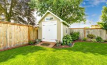 A light brown outdoor shed with white trim and a peaked roof near a wooden fence in the corner of a backyard.