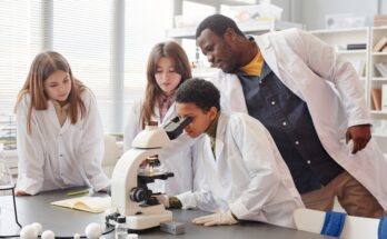 A man and three teenagers wearing white lab coats gather around a microscope set up on a large table.
