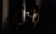 A brown-haired woman in a gray sweatshirt holding a lit candle while sitting next to a window in a dark room.