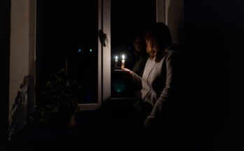 A brown-haired woman in a gray sweatshirt holding a lit candle while sitting next to a window in a dark room.