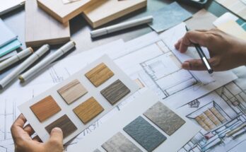 A person holds interior design sketches and material and color samples in one hand and a pencil in the other hand.