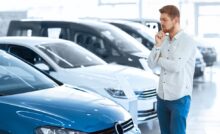 A man is thinking while looking at a blue car with his hand on his chin. He is standing in a car dealership.