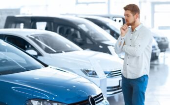 A man is thinking while looking at a blue car with his hand on his chin. He is standing in a car dealership.