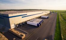 A bird's eye view of an enormous white warehouse building with multiple white semi-trailer trucks parked outside.