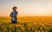 A young man in a blue flannel and a straw hat stands with his arms crossed and smiles in a field of wheat.