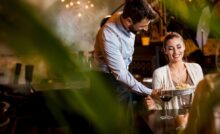 A young woman in a restaurant smiles as a waiter leans over, placing her plate of food in front of her.