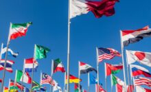 Numerous flagpoles displaying the flags of various nations, including the US, Japan, China, and Russia, beneath a bright blue sky.