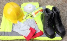 A pile of PPE gear, including a hi-vis vest, gloves, a hard hat, protective glasses, boots, and a respirator.