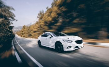 A white car drives down a single-lane road surrounded by trees. The background of the image is blurred by the movement.