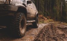 A focus on the side of an off-roading vehicle covered in mud as it drives along a muddy trail on a forested path.