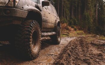 A focus on the side of an off-roading vehicle covered in mud as it drives along a muddy trail on a forested path.