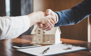 Two people sitting across from each other at a desk shake hands. A pen, documents, and a model house sit on the desk.