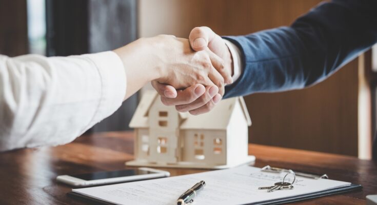 Two people sitting across from each other at a desk shake hands. A pen, documents, and a model house sit on the desk.
