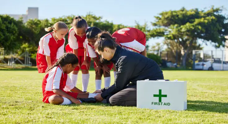 A woman crouching by a group of young girls in sports gear. She is inspecting the leg of one of a girl sitting on the ground.