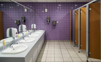Sinks in a public restroom are parallel to stalls. The faucets, soap dispensers, and paper towel dispensers are automatic.