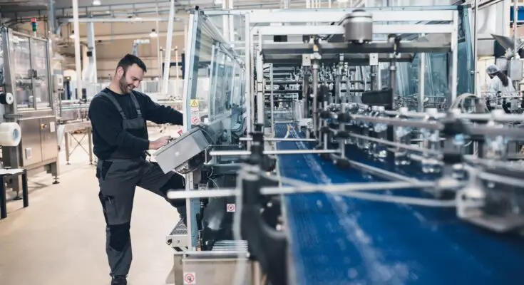 A person operating a touchscreen panel next to an automated conveyor line in a large bottling plant.