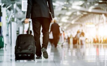 Rear view of a business person walking through an airport with a roller suitcase behind him. Other travelers are around him.
