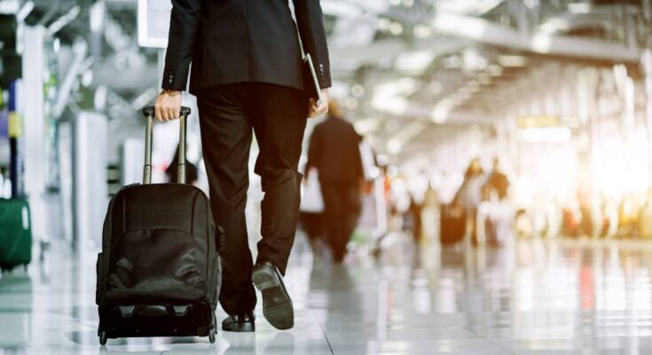Rear view of a business person walking through an airport with a roller suitcase behind him. Other travelers are around him.