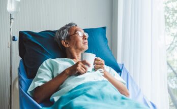 A gray-haired man is lying in a hospital bed with blue linens while holding a white mug and looking out a window.