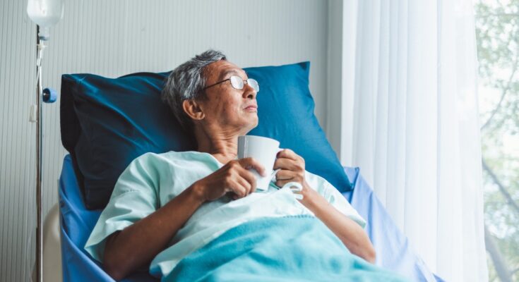A gray-haired man is lying in a hospital bed with blue linens while holding a white mug and looking out a window.
