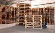 Several stacks of tall wooden pallets in a warehouse. One smaller stack sits on a blue manual forklift.