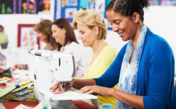 Four women sit next to each other at a table sewing. Each woman is looking down at their sewing machine.