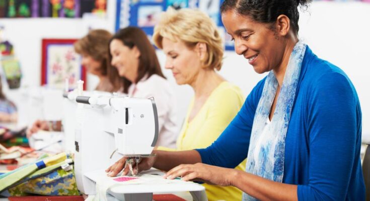 Four women sit next to each other at a table sewing. Each woman is looking down at their sewing machine.
