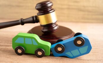 A green toy car and a blue toy car sit in front of a brown gavel on a wooden surface. The blue car is upside down.