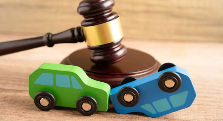 A green toy car and a blue toy car sit in front of a brown gavel on a wooden surface. The blue car is upside down.