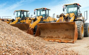 A row of three yellow excavators parked next to each other under a bright blue sky, and in front is a pile of rocks.